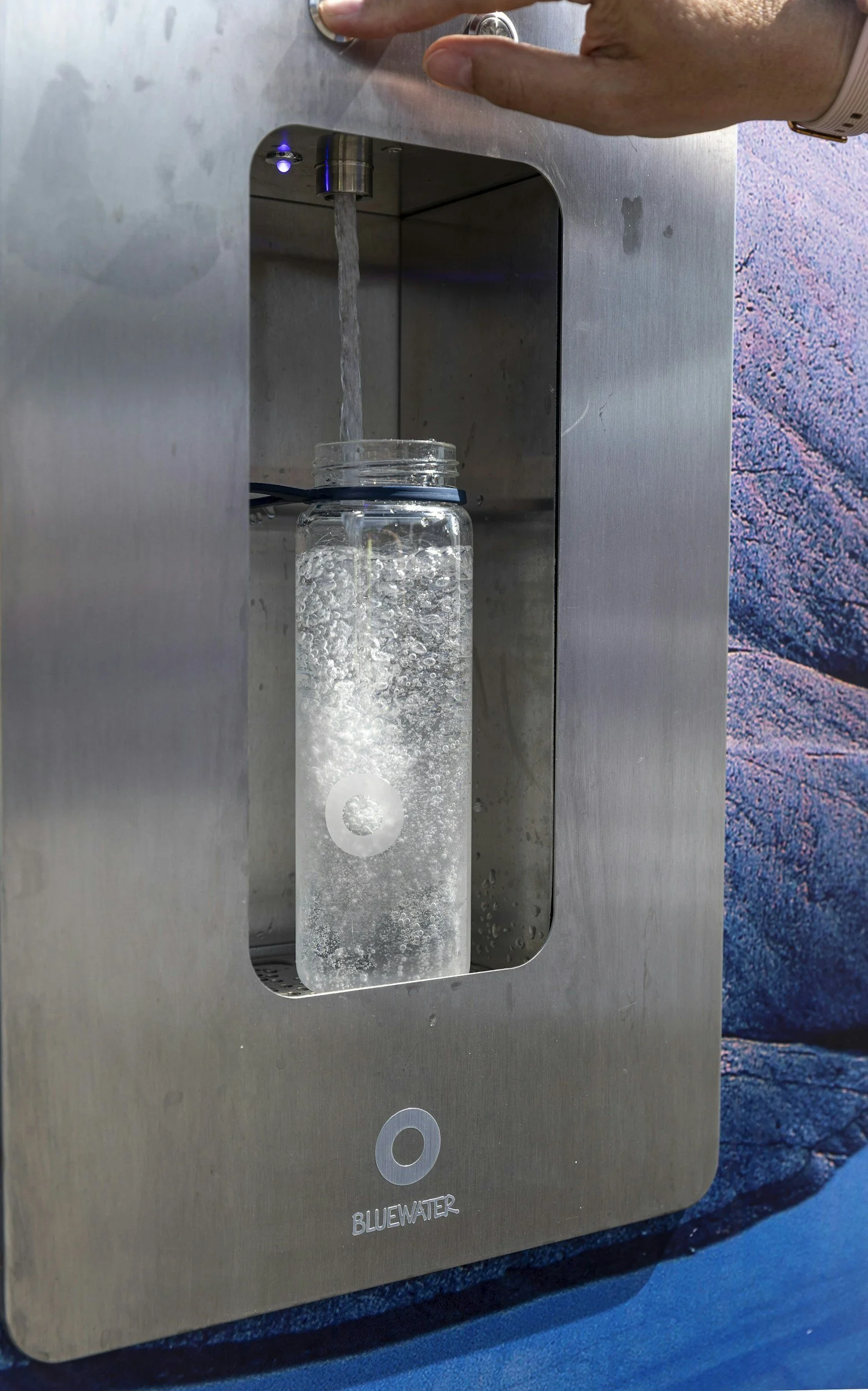 A person pressing a button to dispense water from a stainless steel water fountain. A glass jar is placed underneath the spout with water flowing into it, and bubbles are forming inside the water. The water fountain has a blue light indicator and a circular button, and the brand name 'BLUEWATER' is visible below the button.