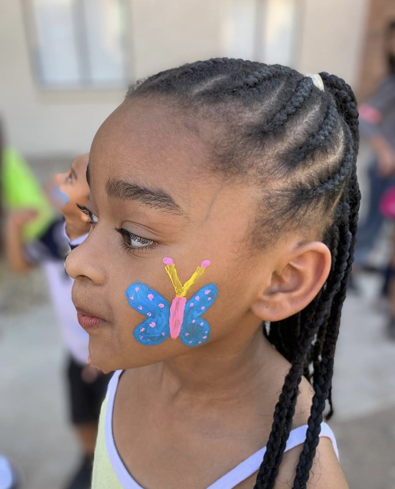 A young girl with braided hair has a colorful butterfly face paint on her cheek. She appears to be at a school or outdoor event, with other children and people blurred in the background.