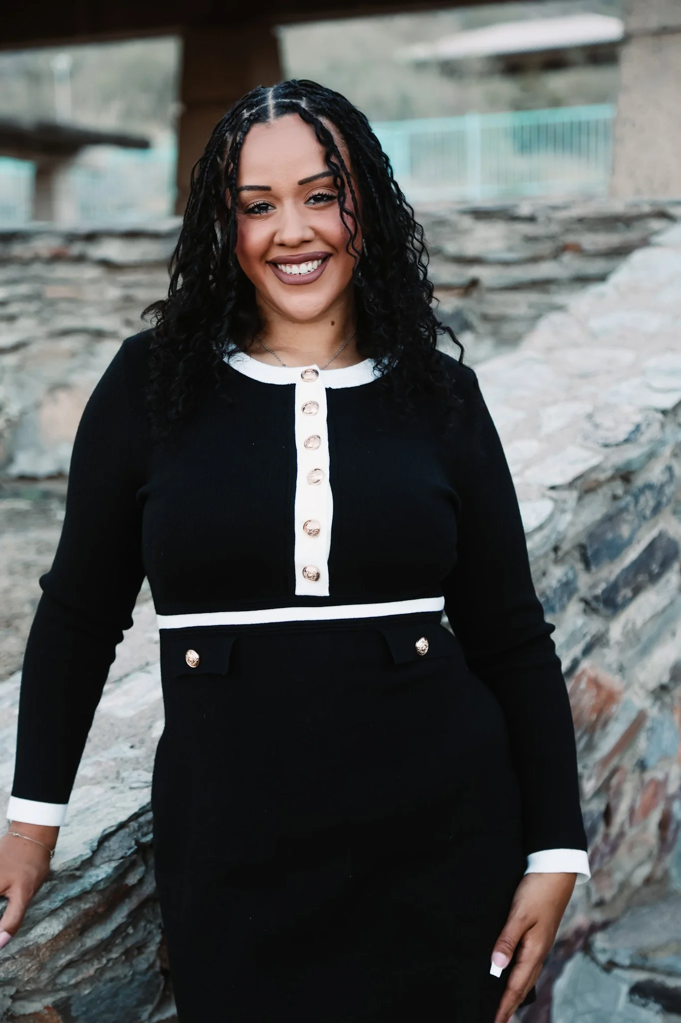 A woman with dark curly hair smiling and wearing a black dress with white accents, standing in front of a stone wall outdoors.