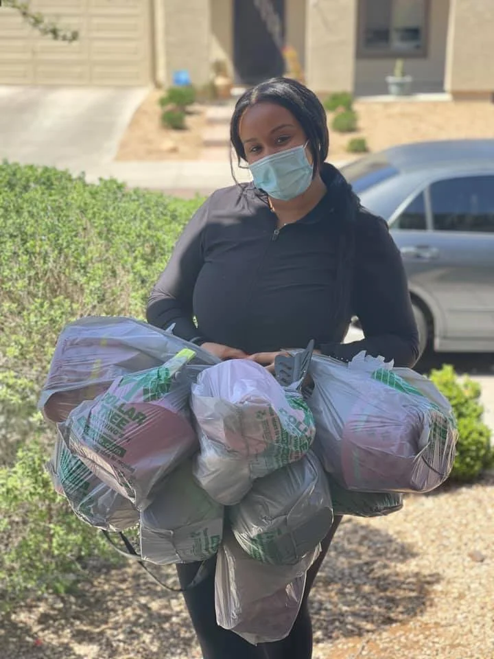 A woman wearing a face mask and black athletic jacket holding grocery bags outside in front of a shrub and a parked car.