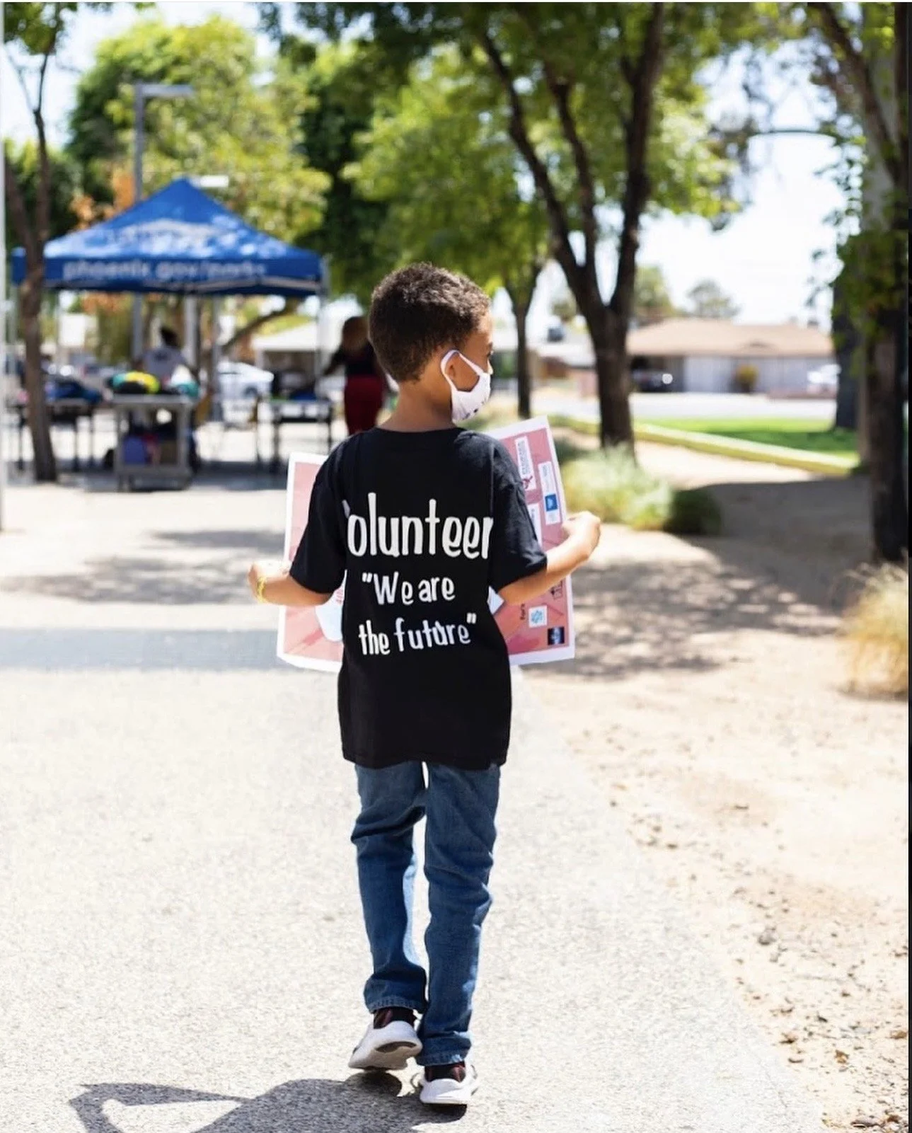 A young boy wearing a face mask, black t-shirt with the words "volunteer 'We are the future'" printed on the back, and blue jeans, walking outdoors on a sunny day and holding a poster board.
