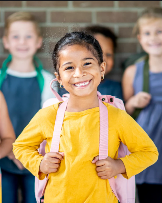 Smiling young girl with a backpack, standing in front of a brick wall, with other children blurred in the background.