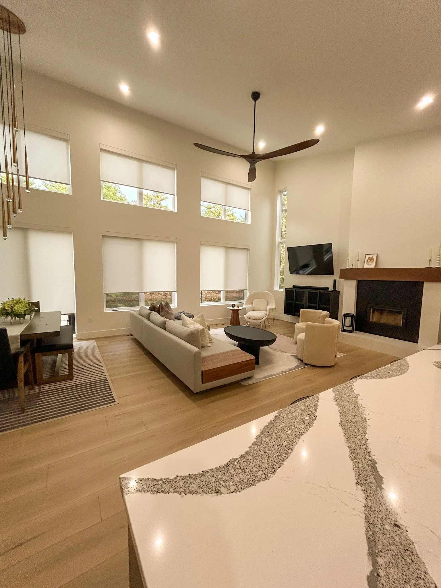 A modern living room with large windows, a ceiling fan, a mounted TV, a fireplace, and beige seating. The room has a light-colored hardwood floor and neutral walls.