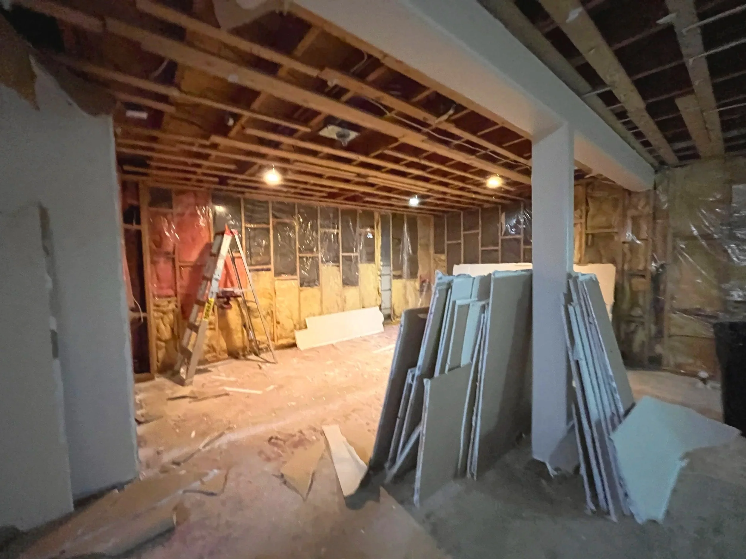Interior of a house under construction with exposed wooden framing, insulation, and partially finished walls, including a ladder and construction materials.