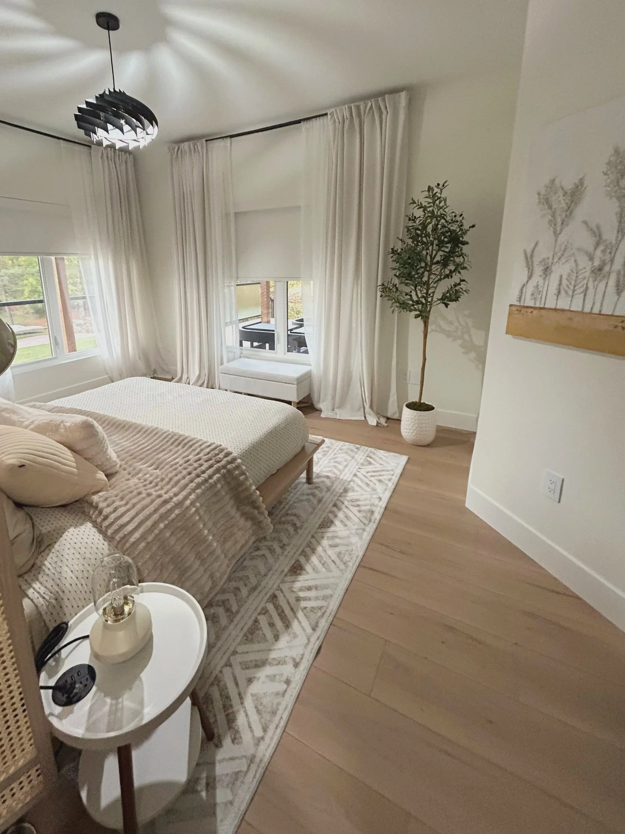 A bedroom with a bed, white curtains, a potted plant, a small white table, and a modern ceiling light, with wooden flooring and a view of a balcony.