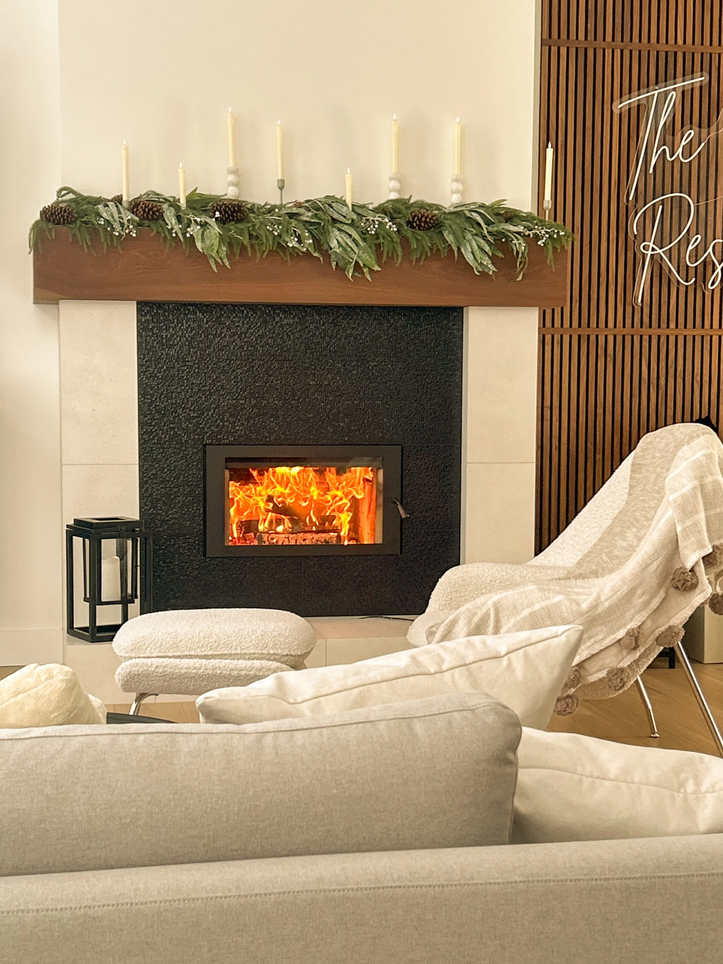 Living room with a beige sofa, a small matching beige ottoman, a cream throw blanket, and a modern armchair. A fireplace with an orange glowing fire is centered on the wall, decorated with a wooden mantel adorned with green foliage and pinecones, with white candles. To the right, a decorative sign with cursive writing reads 'The Res' on a wooden slat wall.
