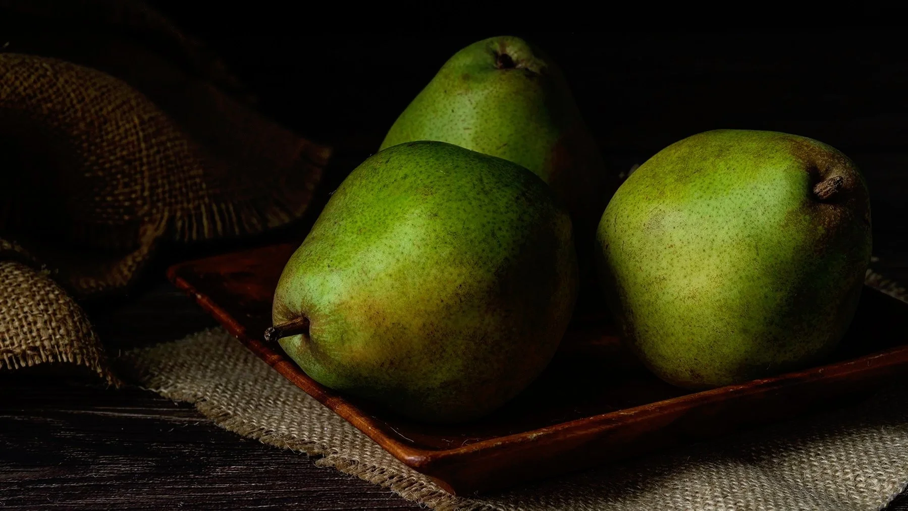 Three green pears on a wooden tray, with burlap fabric and dark wooden surface in the background.