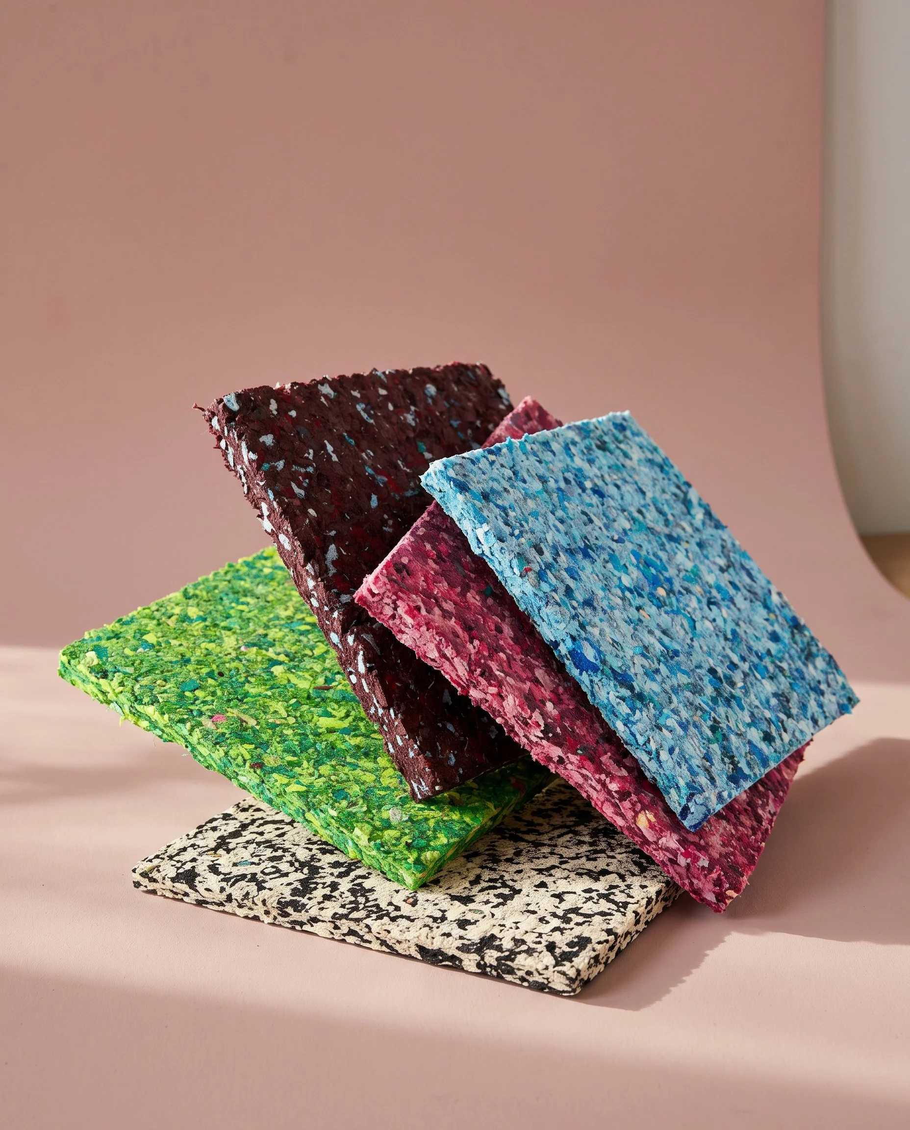 Stacked colorful architectural tiles, including green, black and white, pink, blue, red, and dark colors, on a soft pink background.