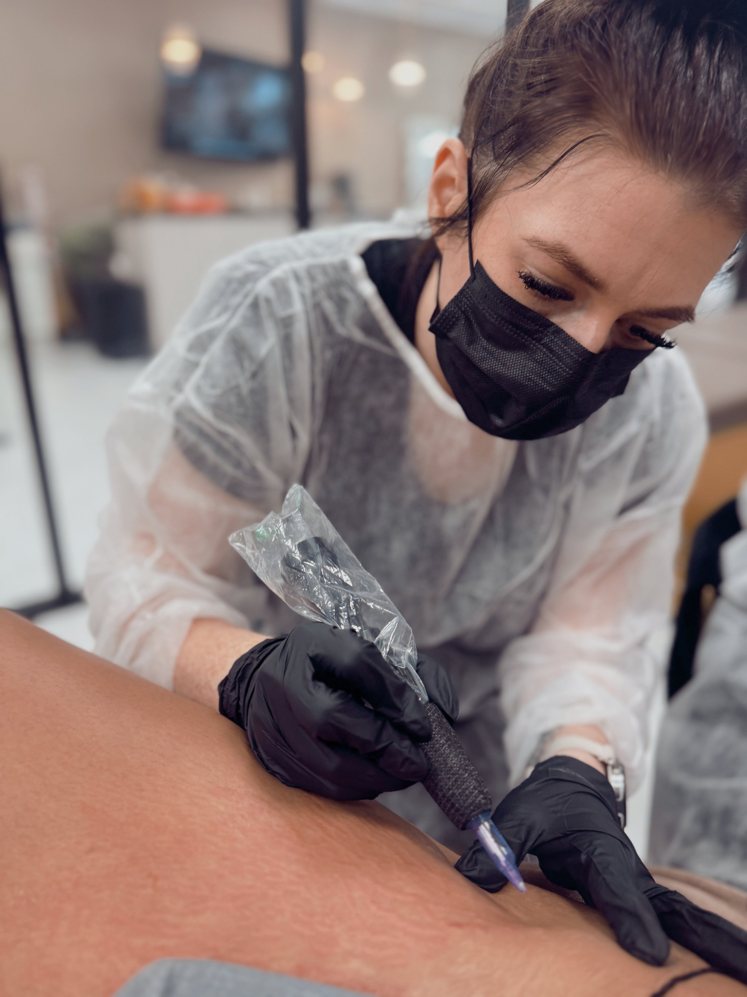 Close-up of a person receiving a tattoo, with a tattoo artist wearing black gloves and a face mask, working on the person's arm in a studio.
