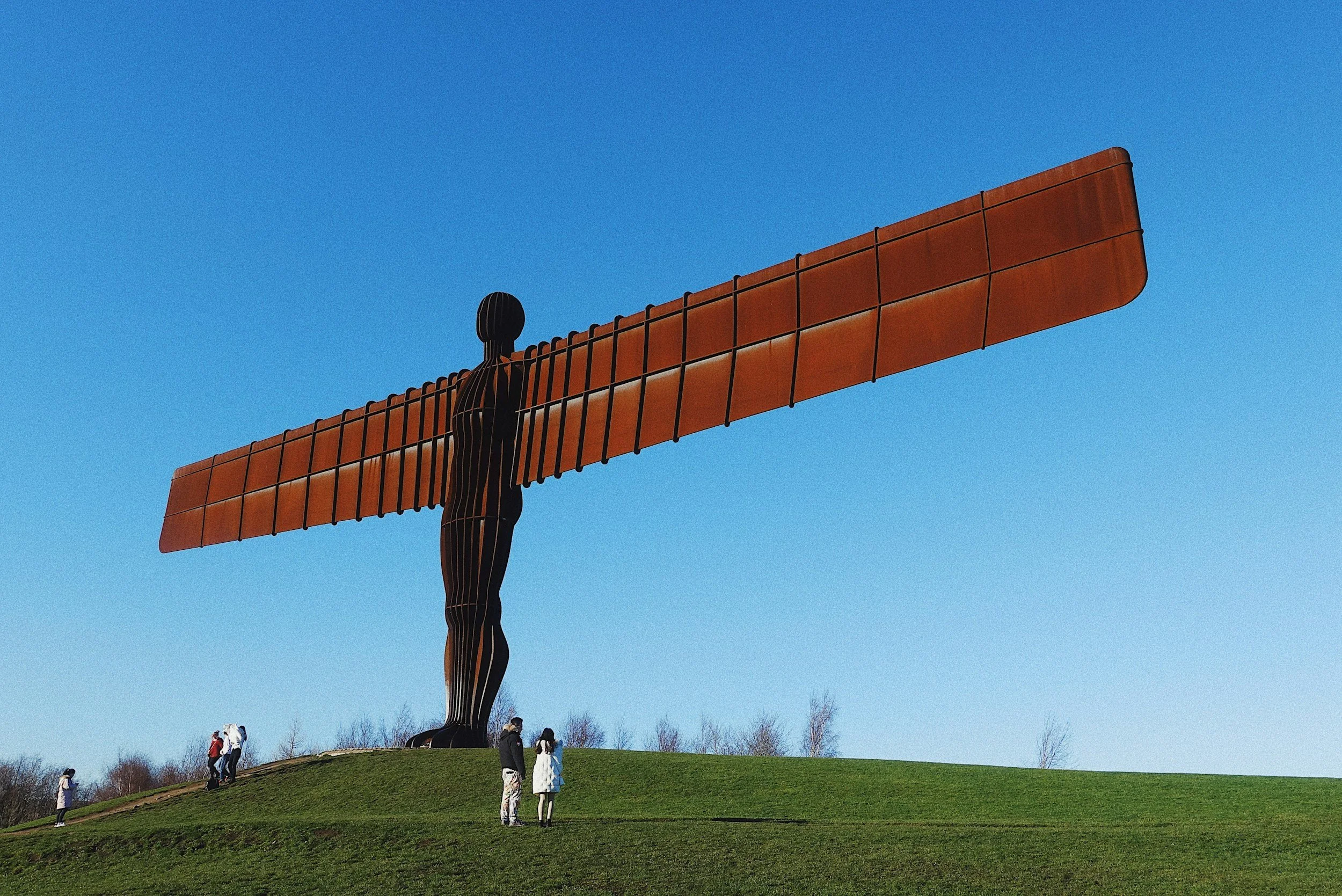 A large statue of a woman holding a big orange windmill with a blue sky background. Several people are standing and walking around the grassy hill at the base of the statue.