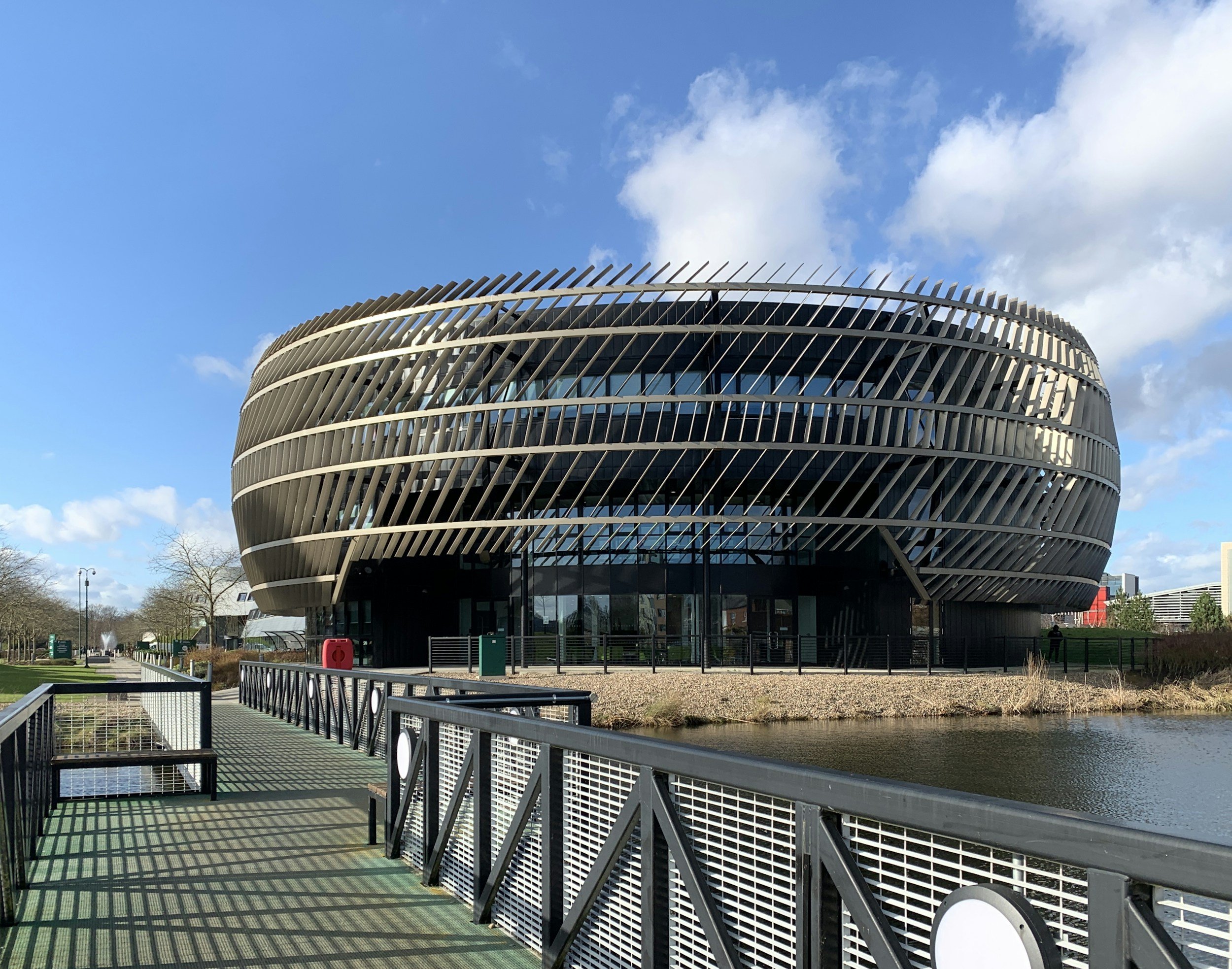 Modern building with curved architecture and horizontal slats, surrounded by a canal, walkways, and a park with trees under a partly cloudy sky.