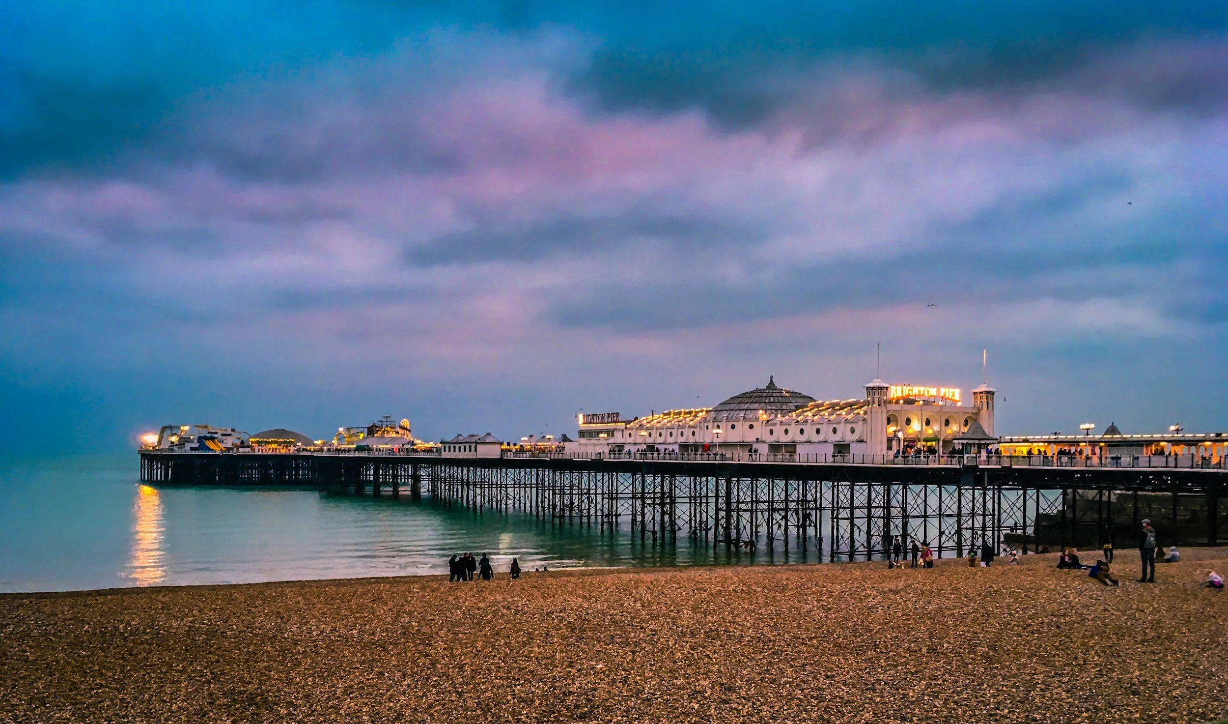 Brighton Pier at dusk with a cloudy sky and people walking on the beach in the foreground.