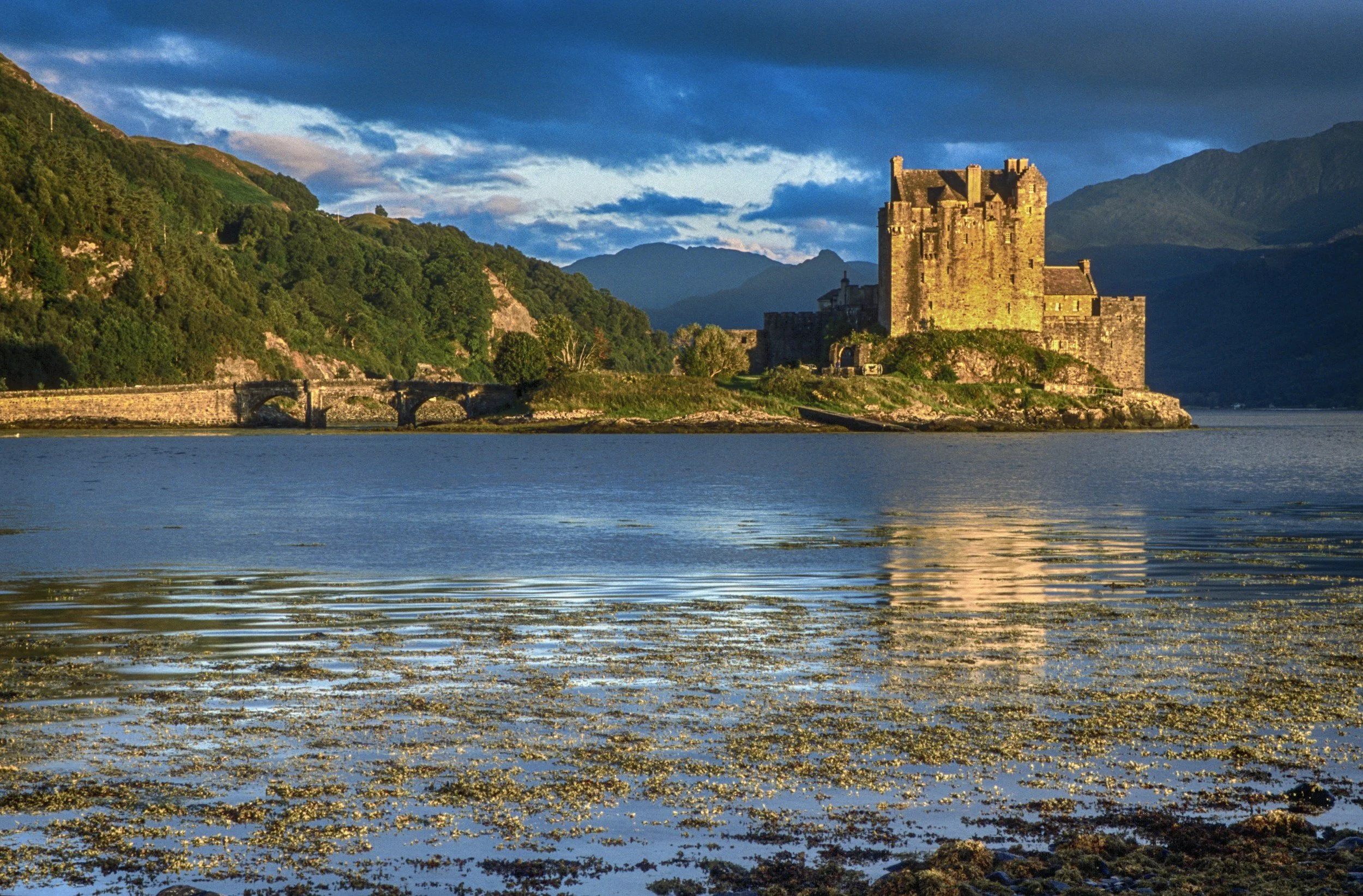 A castle on the shore with a stone bridge, surrounded by green hills, under a cloudy sky during daytime.