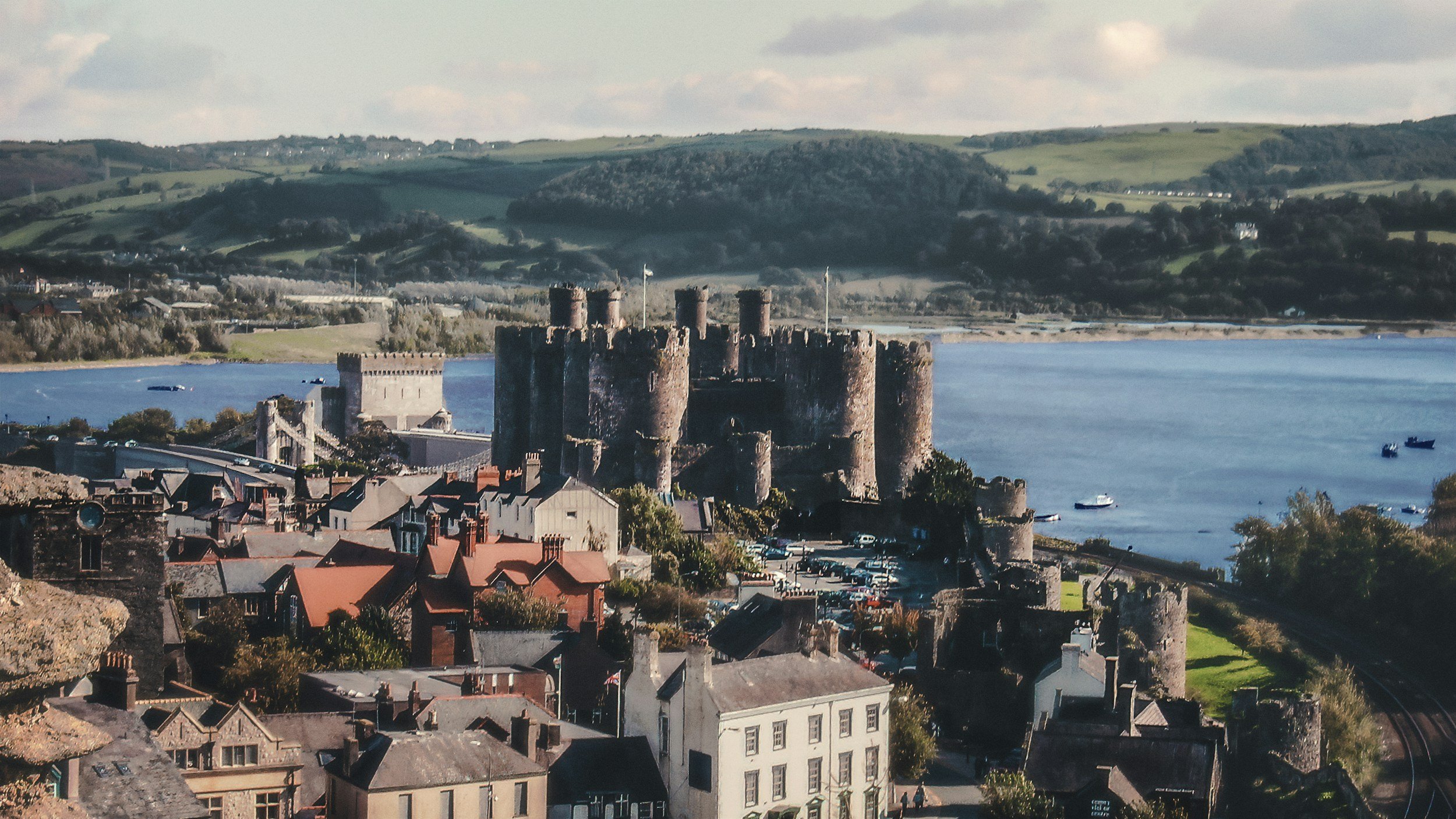 A historic castle situated on a riverbank with boats sailing nearby, surrounded by a town with residential buildings and green rolling hills in the background.