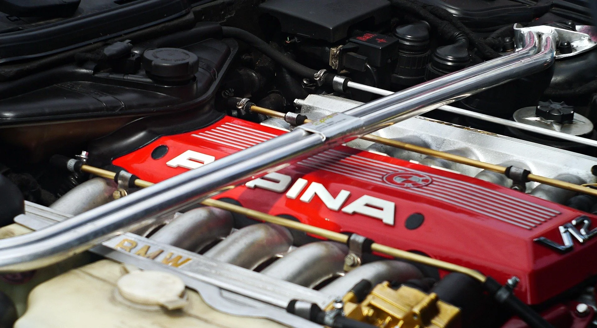 Close-up of a car engine with a red valve cover labeled 'DINA' and various metallic components, hoses, and black plastic parts.