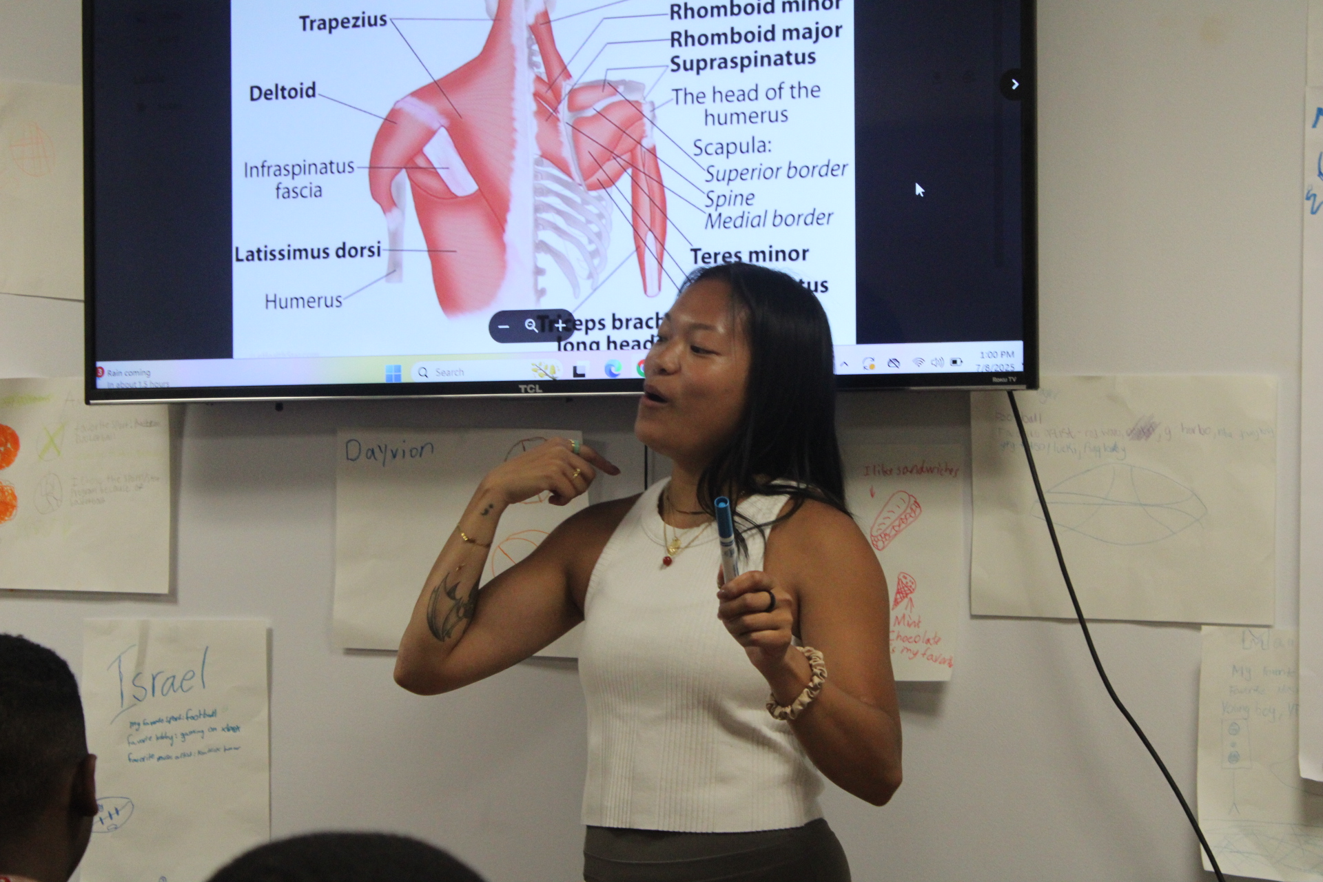 Teacher pointing to a shoulder muscle anatomy diagram on a large screen.