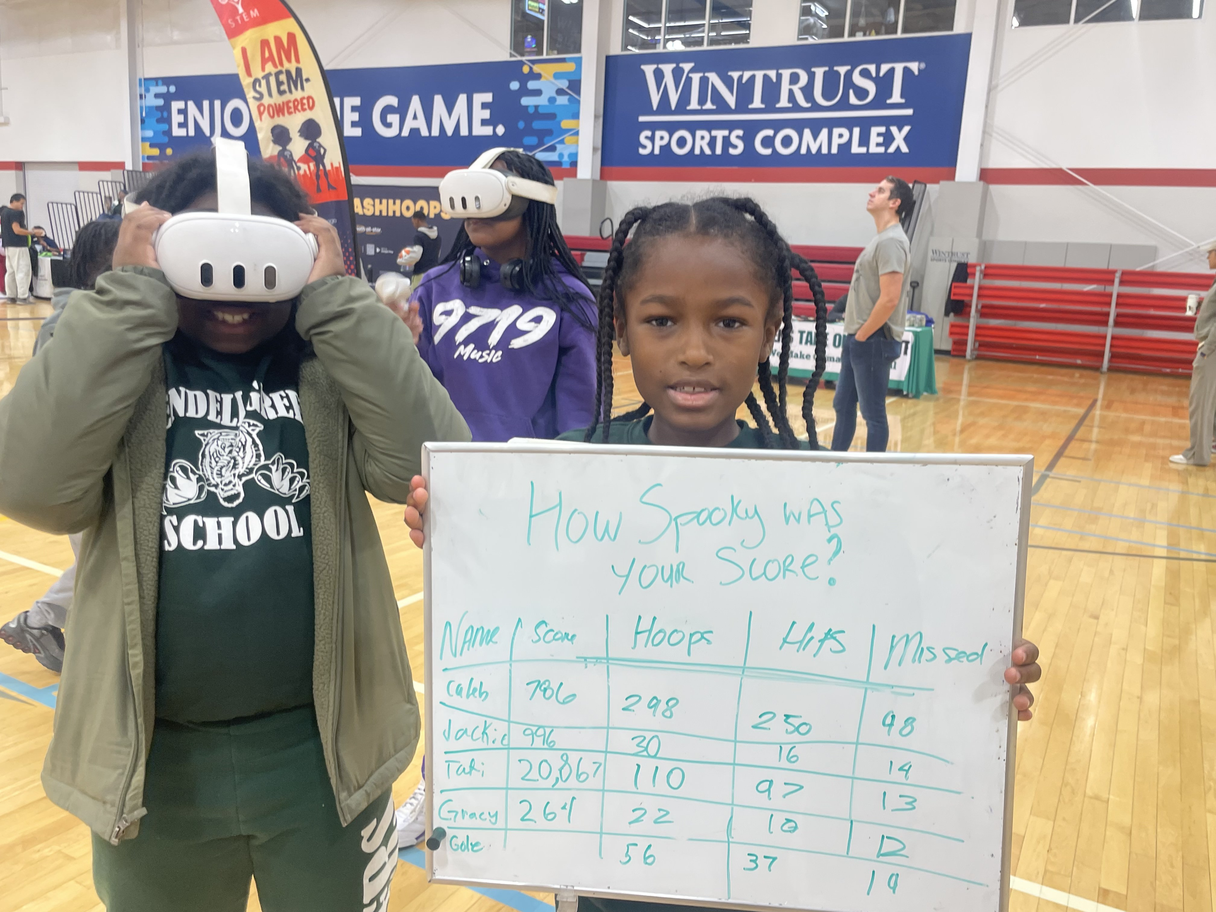 Child holding scoreboard while two peers wear VR headsets at sports event.
