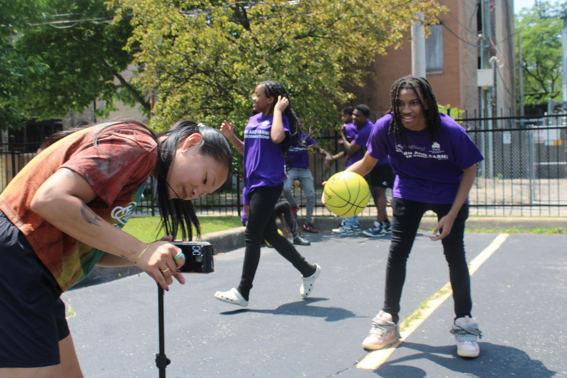 Person filming kids in purple shirts playing basketball outdoors.