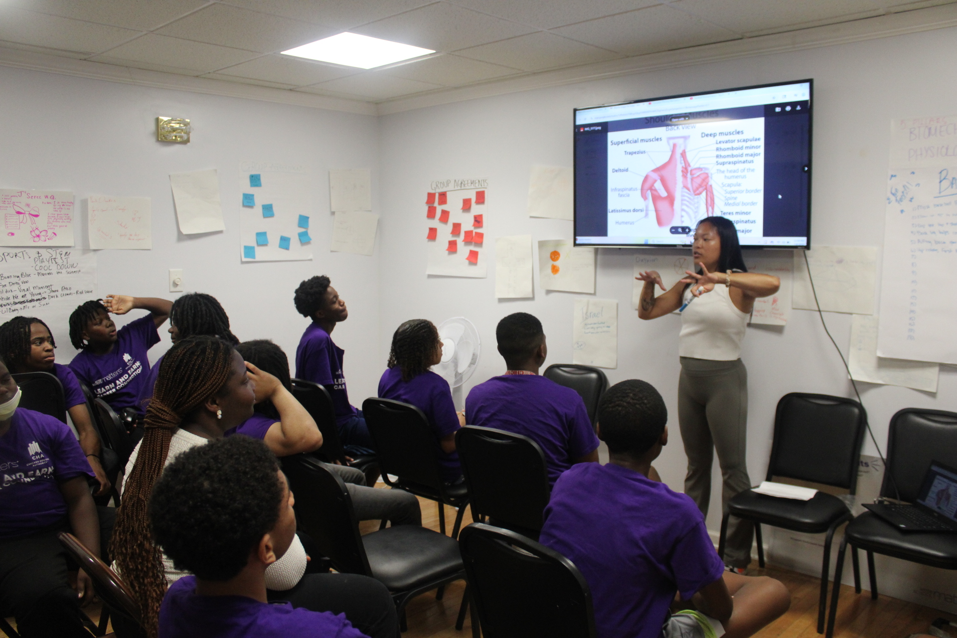 Teacher instructing youth in purple shirts using a muscle anatomy display.