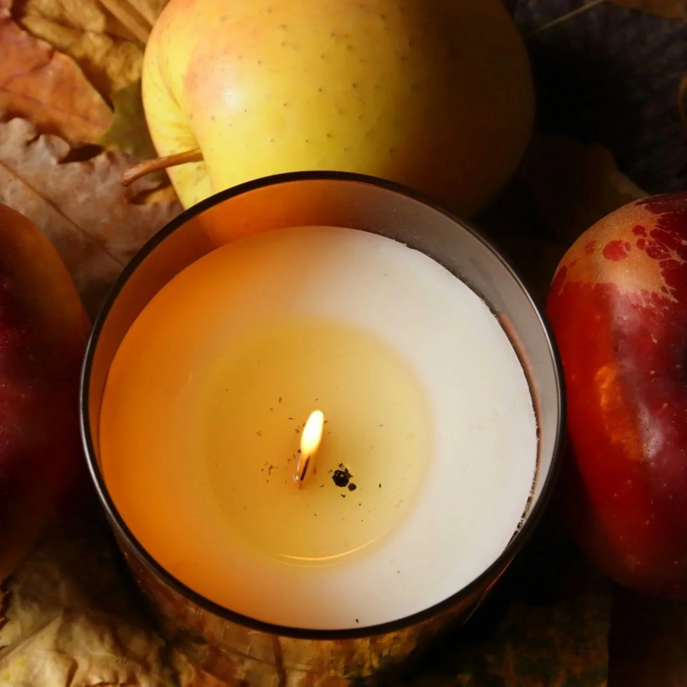 A lit candle in a metallic holder surrounded by apples and dried leaves.