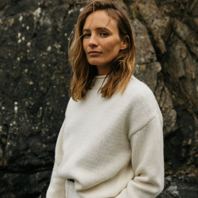 Woman with shoulder-length brown hair wearing a white sweater standing outdoors near rocks.