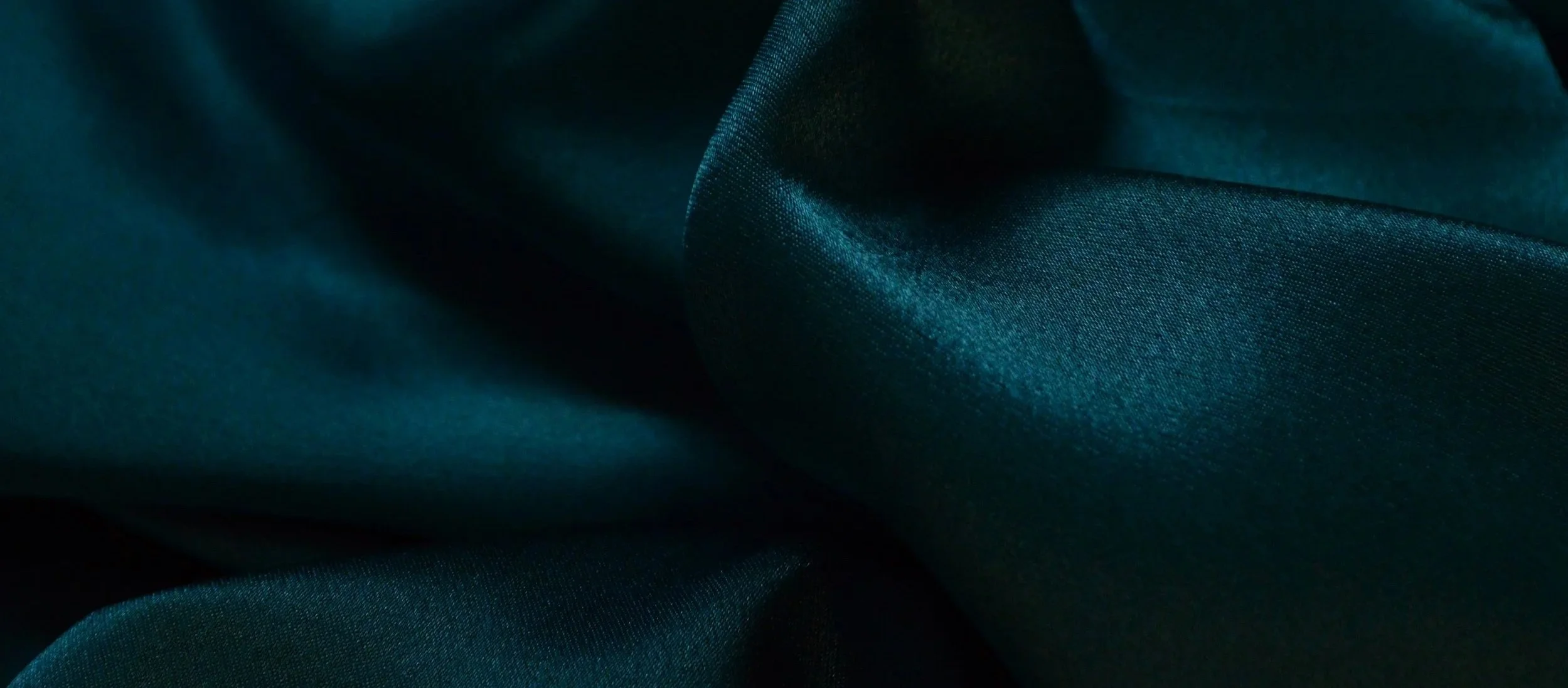 Close-up of dark teal silk fabric with smooth, flowing folds and a subtle sheen.