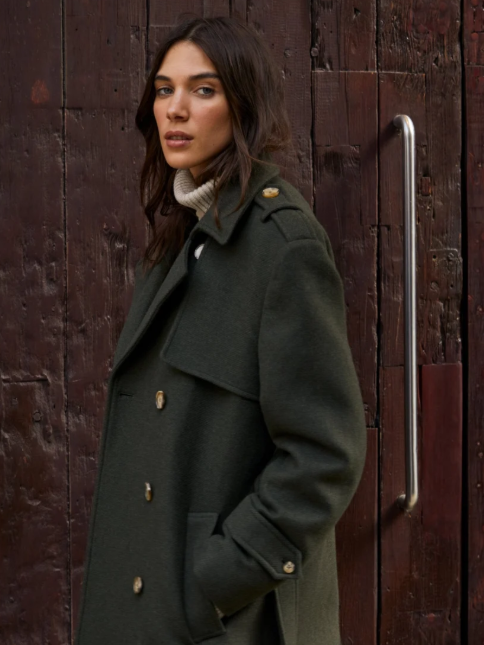 Woman with dark brown hair wearing a dark green military-style coat with gold buttons, standing against a wooden background.