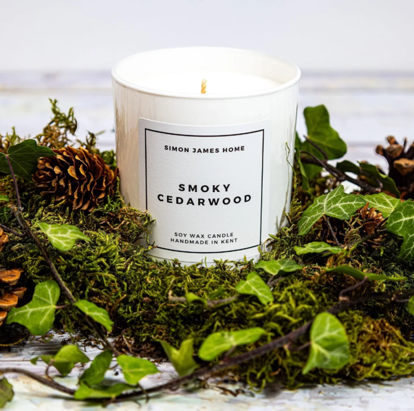 White soy wax candle labeled 'Smoky Cedarwood' surrounded by moss, ivy, and pinecones.
