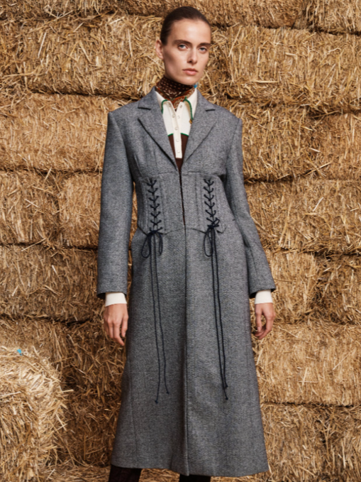 Fashion model wearing a long gray coat with lace-up details standing in front of hay bales.