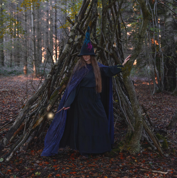 Person dressed as a witch in a dark cloak and hat standing in a woodland clearing with a makeshift tent made of branches and sticks.