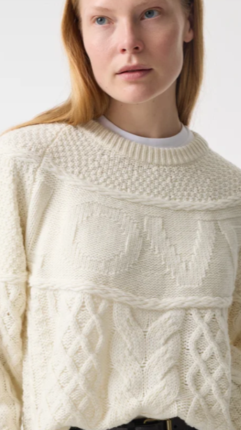 Close-up of a woman with fair skin and red hair wearing a cream-colored cable knit sweater with textured patterns.