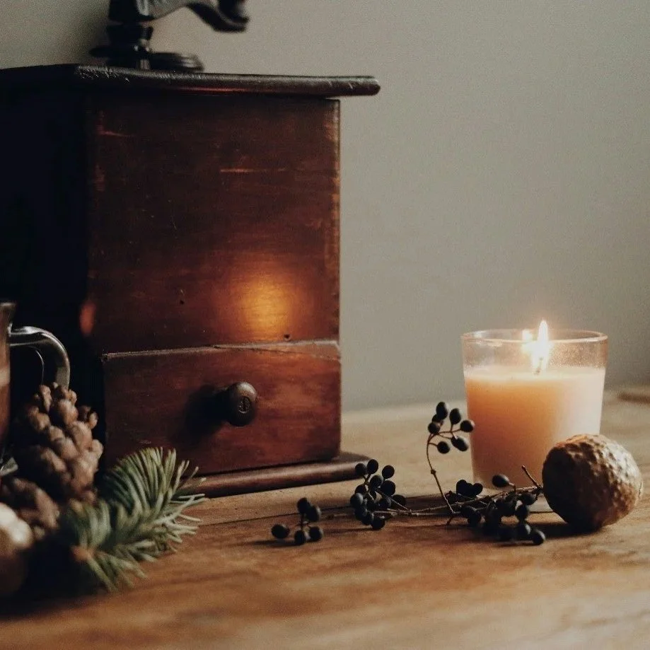 Close-up of a wooden table with a lit candle, sprigs of black berries, pinecone, and evergreen branch, with a dark wooden box nearby.