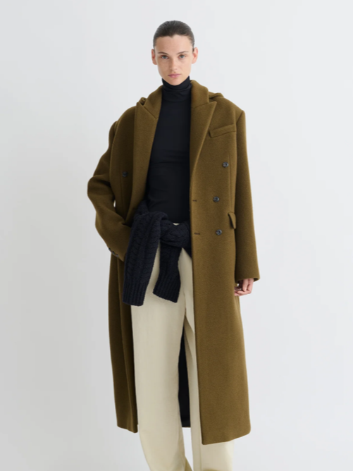 Woman dressed in a black turtleneck, beige pants, and an oversized olive green coat, with a dark knit sweater tied around her waist, standing against a plain white background.