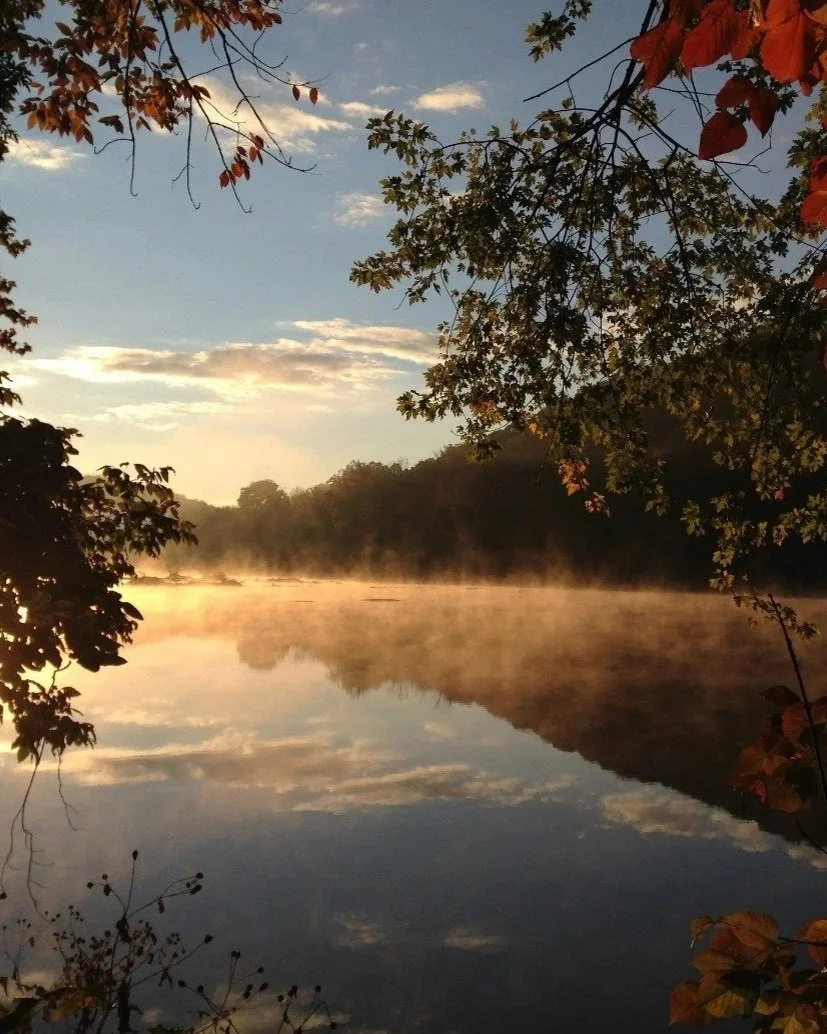 A calm river with mist rising from the water in the early morning or late evening, surrounded by trees with autumn-colored leaves, under a partly cloudy sky.