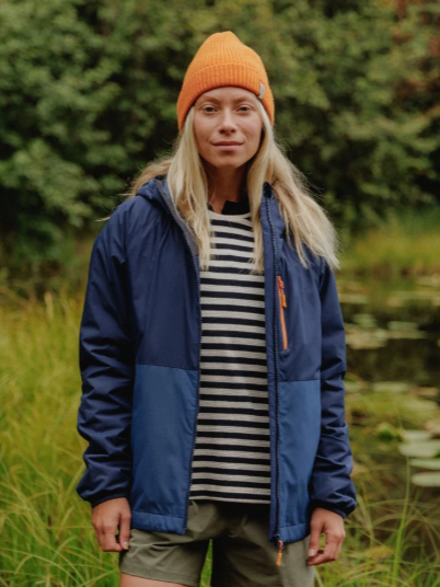 A woman standing outdoors near a pond with green grass and trees in the background, wearing a blue jacket, striped shirt, orange beanie, and khaki shorts.