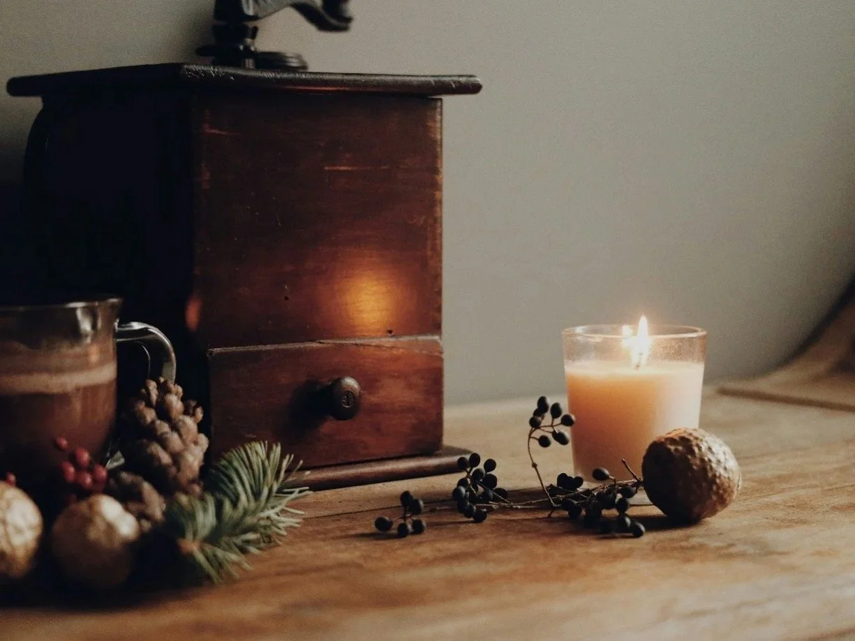A lit candle, a cookie, and black berry-like fruits on a wooden table, with a small wooden drawer or box, a coffee cup, and some pine cones and greenery in the background.
