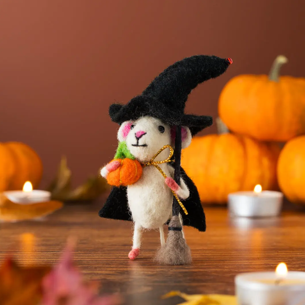 Felted witch mouse holding a pumpkin, standing on a wooden surface with pumpkins and tea light candles in the background.