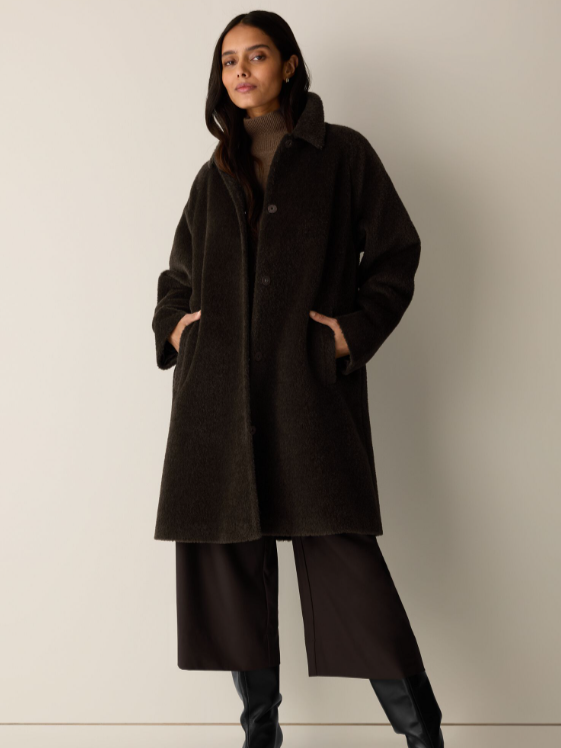 Woman standing against a plain light-colored wall, wearing a dark wool coat, wide-leg black pants, and black boots.