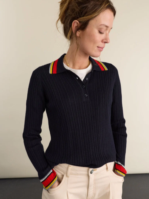 Woman in a black ribbed sweater with colorful striped collar and cuffs, beige pants, standing with hands in pockets, looking downward.