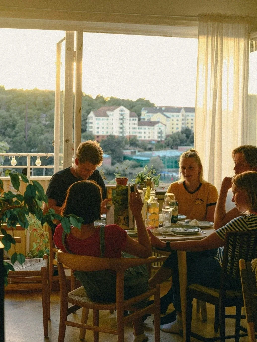 A group of five friends sitting around a dining table enjoying a meal and beverage, with a large window showing a sunset view of a hillside with apartment buildings in the background.