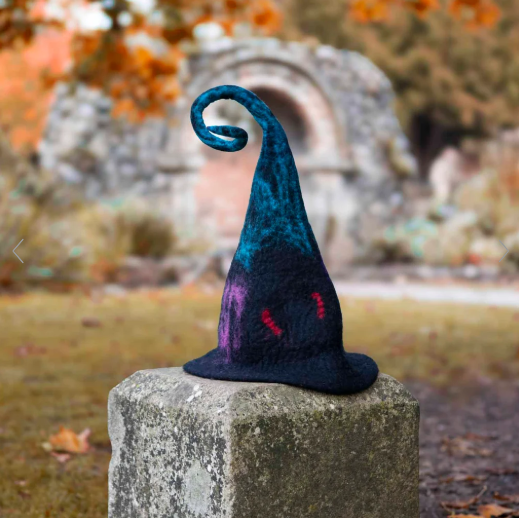 Colorful, felted wizard hat with a long, curled tip rests on a stone pedestal outdoors with autumn trees in the background.