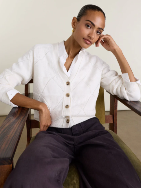 A woman sitting in a vintage wooden armchair with a green cushion, wearing a white sweater with buttons and black pants, posing with her hand near her face.