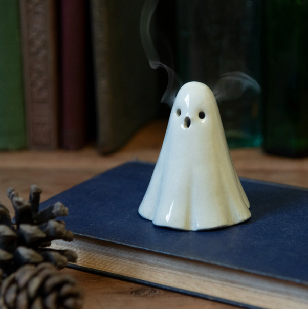 A ceramic ghost-shaped incense holder with smoke rising from the top, placed on a closed book on a wooden surface.