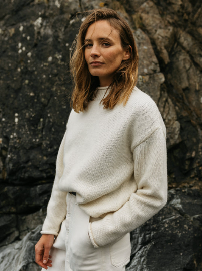 A woman with shoulder-length brown hair wearing a white sweater and light-colored pants standing outdoors near rocky terrain.