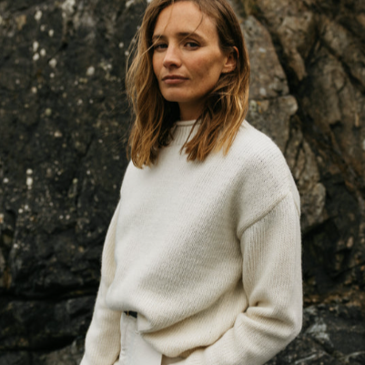 Woman with wavy hair wearing a white sweater standing outdoors near rocks.
