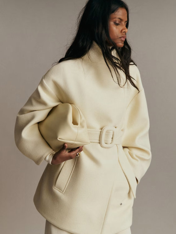 Woman in a cream-colored oversized coat with a belt, standing sideways with her eyes closed.