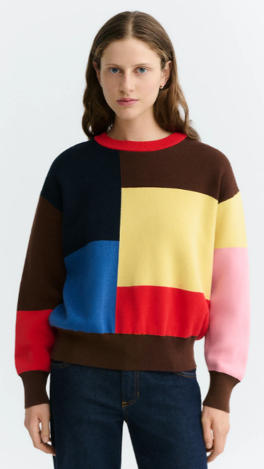 Young woman with long brown hair wearing a colorful patchwork sweater and blue jeans standing against a plain white background.