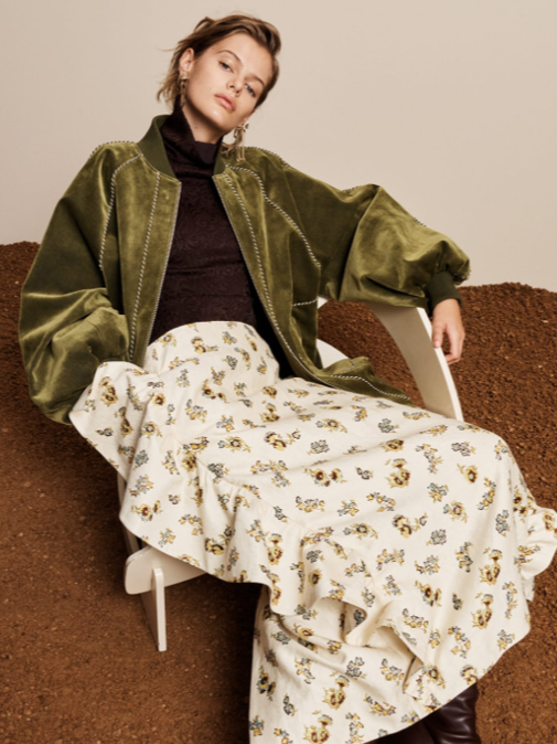 A young woman with short hair sitting on a white chair, wearing a green bomber jacket, black top, and a long floral skirt