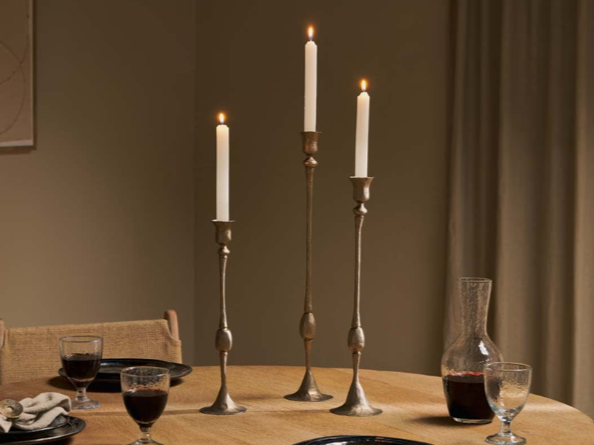 A dining table with three tall white candles in brass candle holders, lit and burning steadily. The table has glasses of red wine, a decanter, plates, and utensils, with a beige wall and curtains in the background.