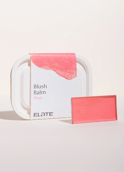 A pink blush balm in a rectangular shape, next to its packaging box labeled "Blush Balm Pinch" with a large pink swatch on it, placed in front of a white tray.